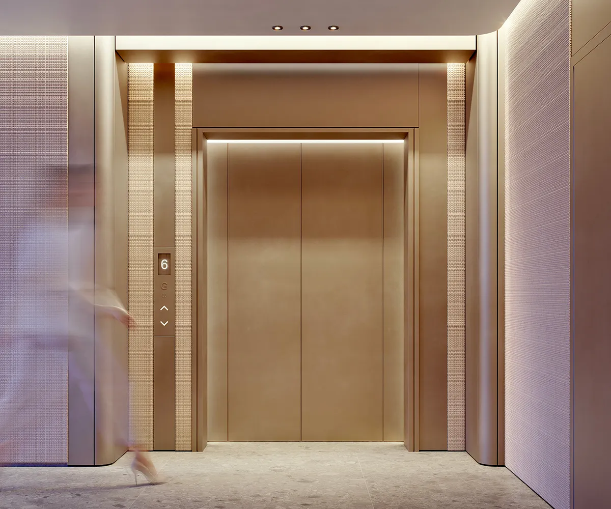 Imagen ilustrativa de un vestíbulo de ascensores: puertas de bronce cepillado entre paneles texturizados y una figura que pasa con efecto de movimiento.